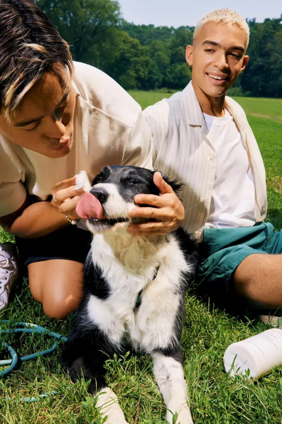 Two individuals sit on grass in a park; the person on the left wipes the face of a black and white dog while the other person sits beside them smiling.