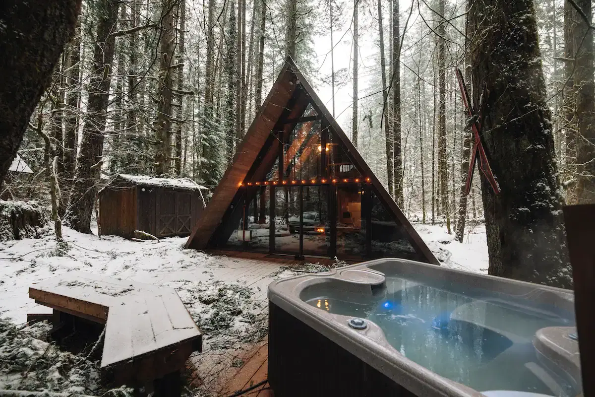 A snowy forest surrounds an A-frame cabin with string lights and a steaming hot tub on a wooden deck.