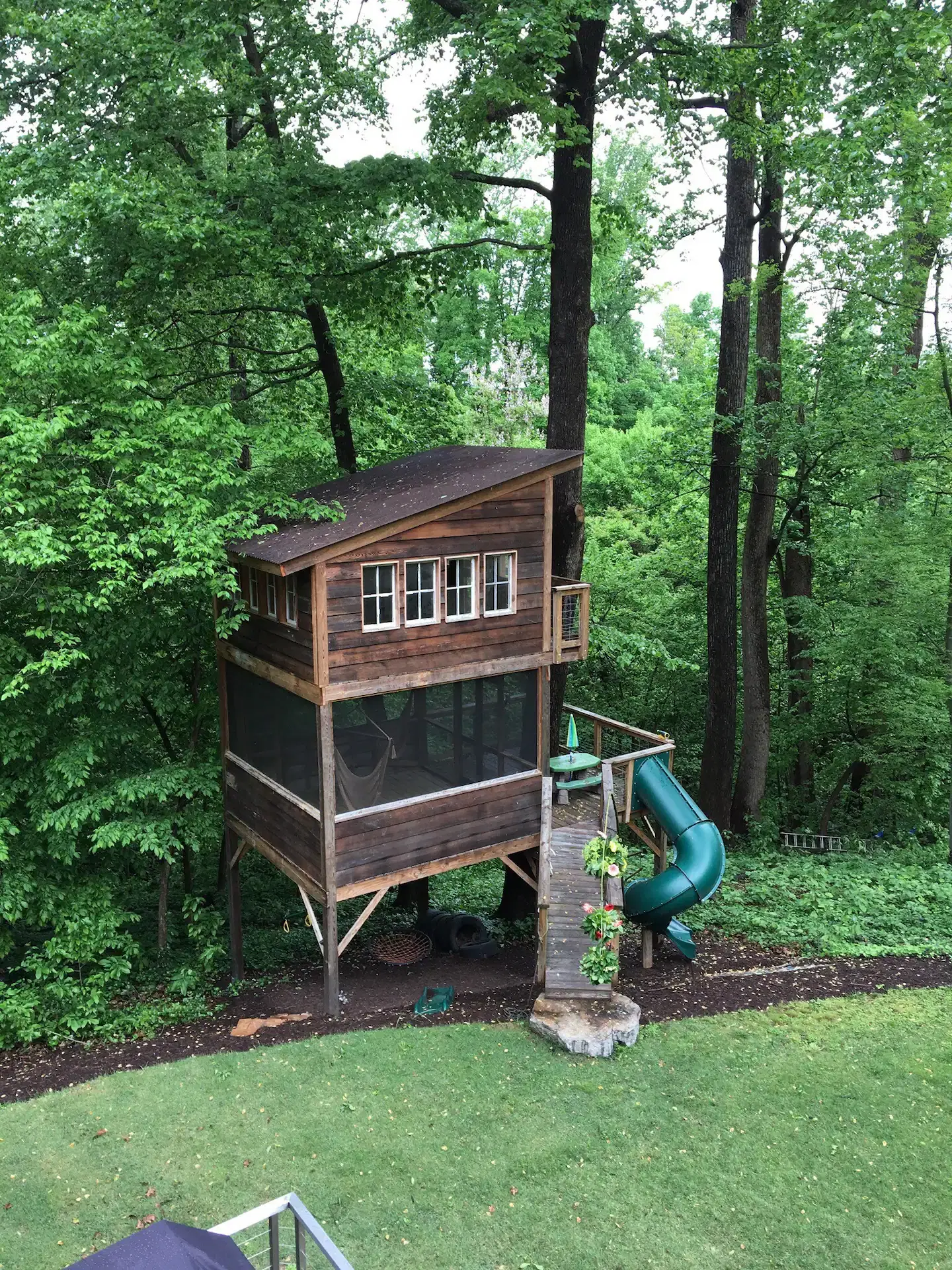 A wooden treehouse on stilts stands in a lush forested area with a slide attached.