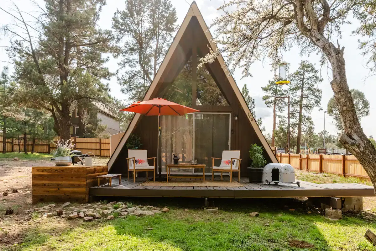 An A-frame cabin with a front deck nestled in a yard with trees and a wooden fence.