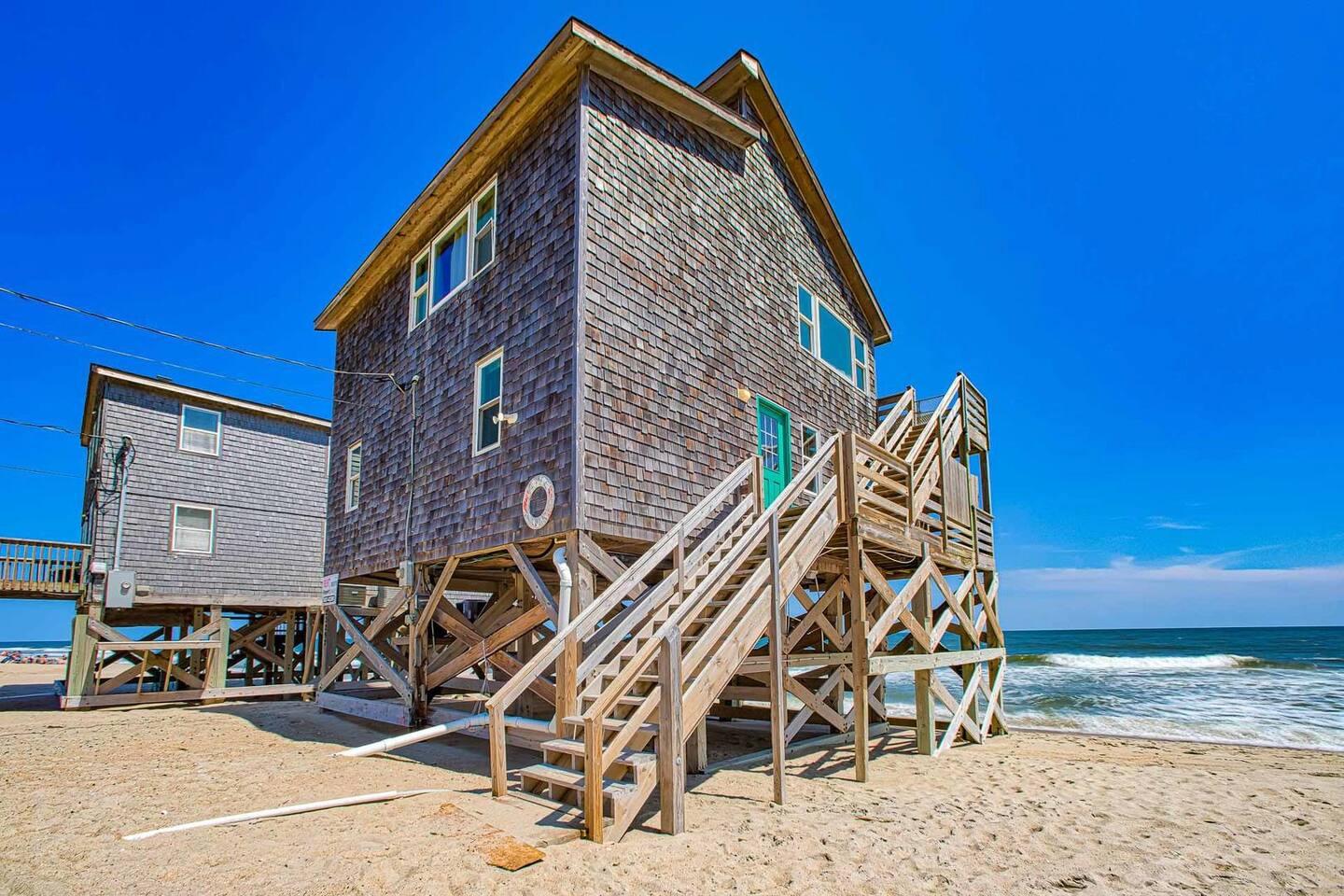 A wooden beachfront house on stilts has a staircase leading up to a porch.