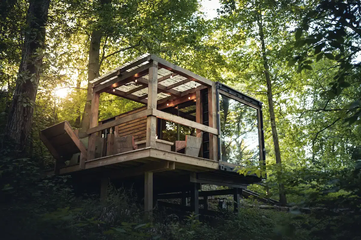 A small wooden cabin with large glass panels stands elevated among lush green trees.