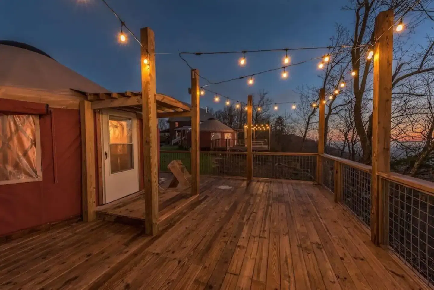 A wooden deck with string lights at dusk features a small structure with a white door and trees in the background.