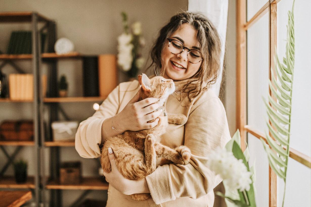 A woman with glasses smiles while petting an orange cat near a window.