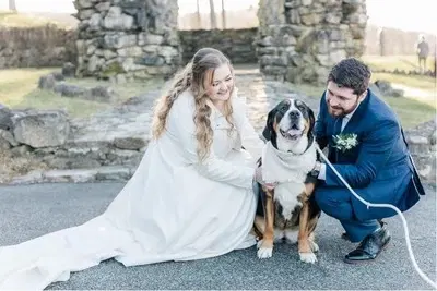 A bride and groom kneel beside a large dog wearing a scarf in front of stone ruins.