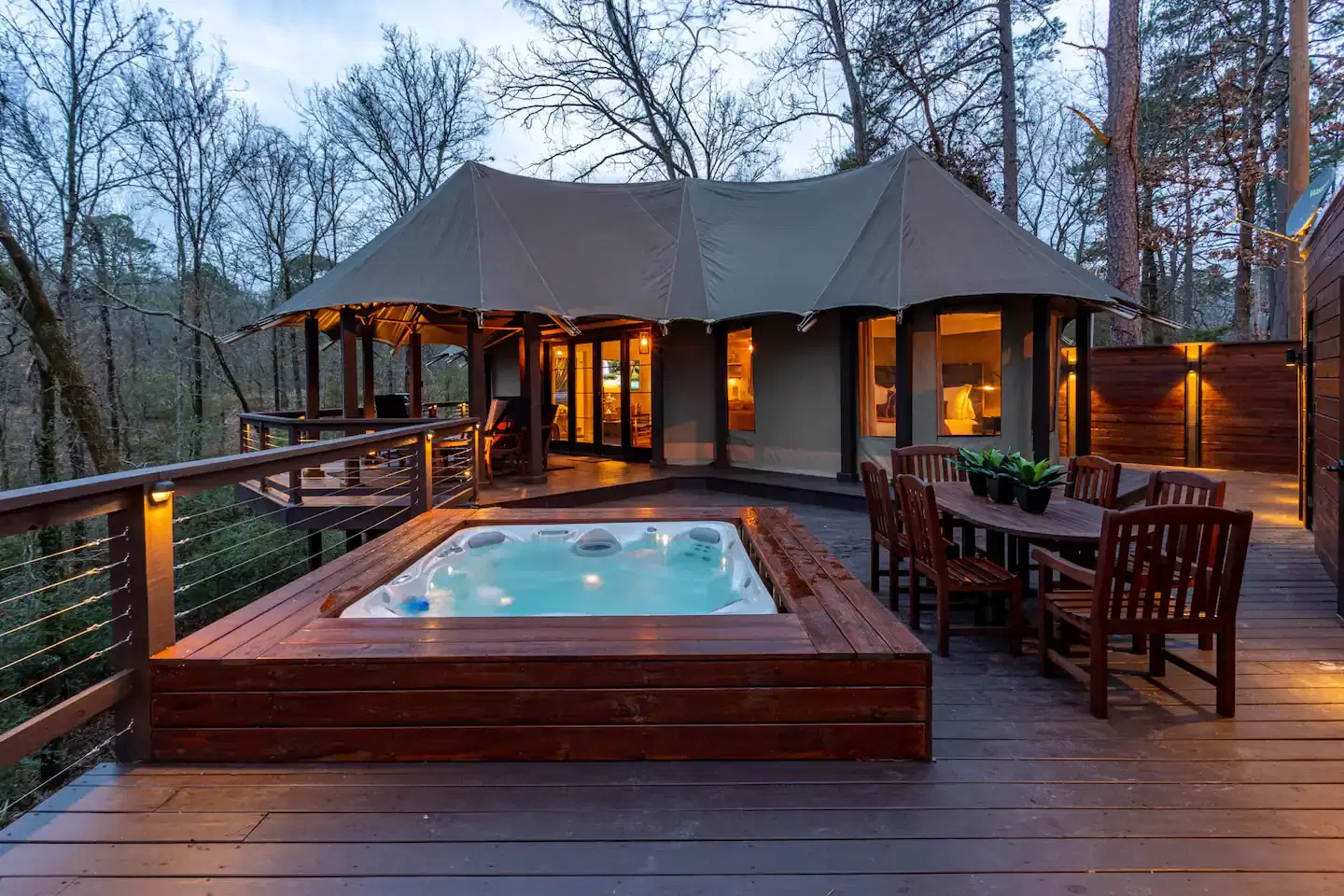 A modern glamping tent with a hot tub on a wooden deck surrounded by trees at dusk.