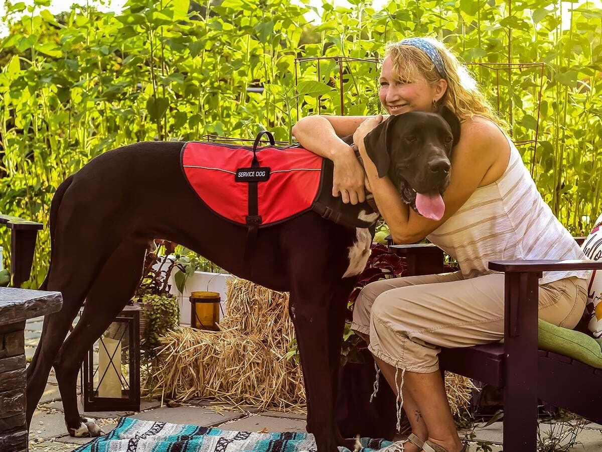 A woman hugs a large service dog in a red vest outside.