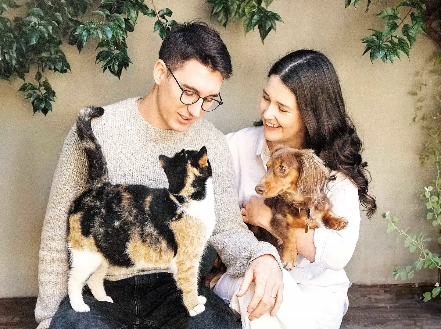 A smiling man with a calico cat on his lap sits with a woman holding a small brown dog against a backdrop of greenery.