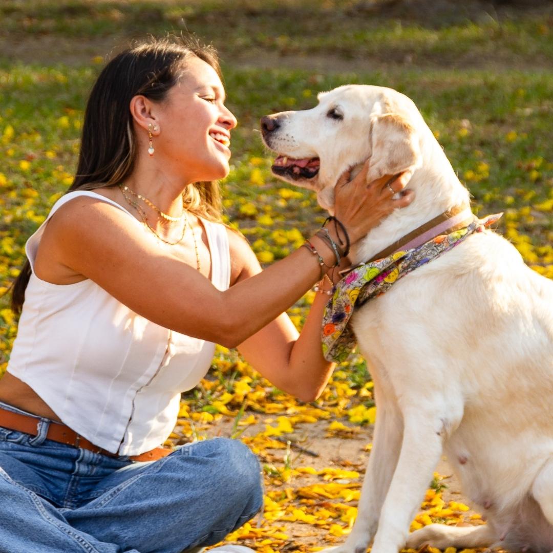 A woman sitting on the grass smiles and lovingly holds a yellow Labrador retriever wearing a colorful bandana. They are outdoors on a sunny day with yellow petals scattered on the ground.