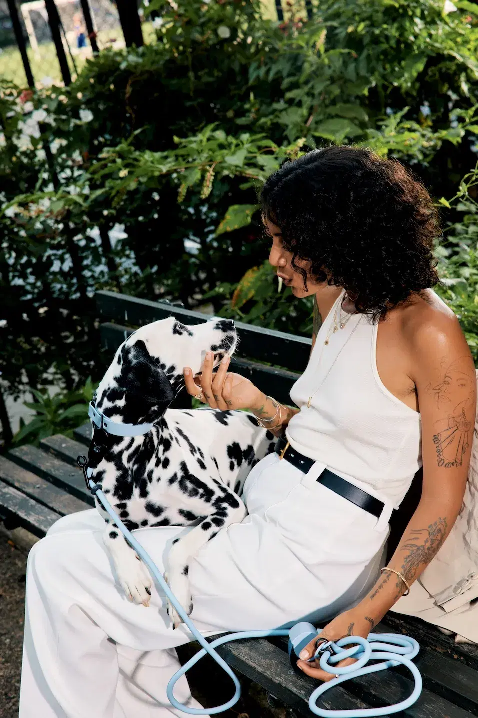 A woman with curly hair and tattoos sits on a bench petting a Dalmatian dog.
